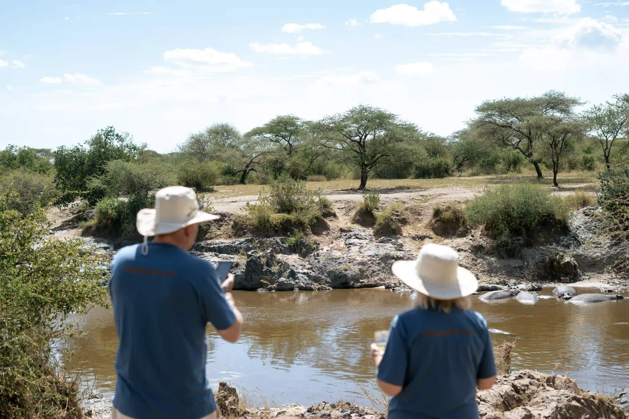 Two guests on a riverbank watching hippos lying in the brown water